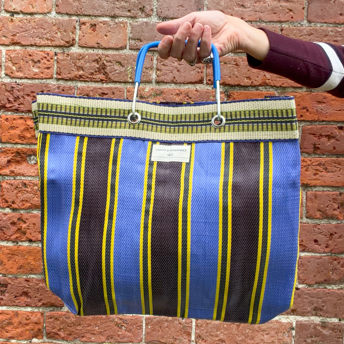 A hand holding a striped woven tote bag with blue handles, set against a red brick wall background. The bag features vertical stripes in blue, dark brown, and yellow, with a cream-colored brand label that reads 'The Bag Company'.