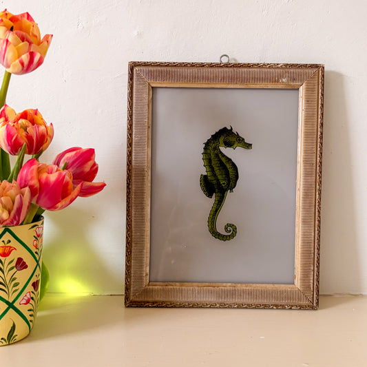 Framed artwork of a seahorse next to a vase with tulips on a light surface.