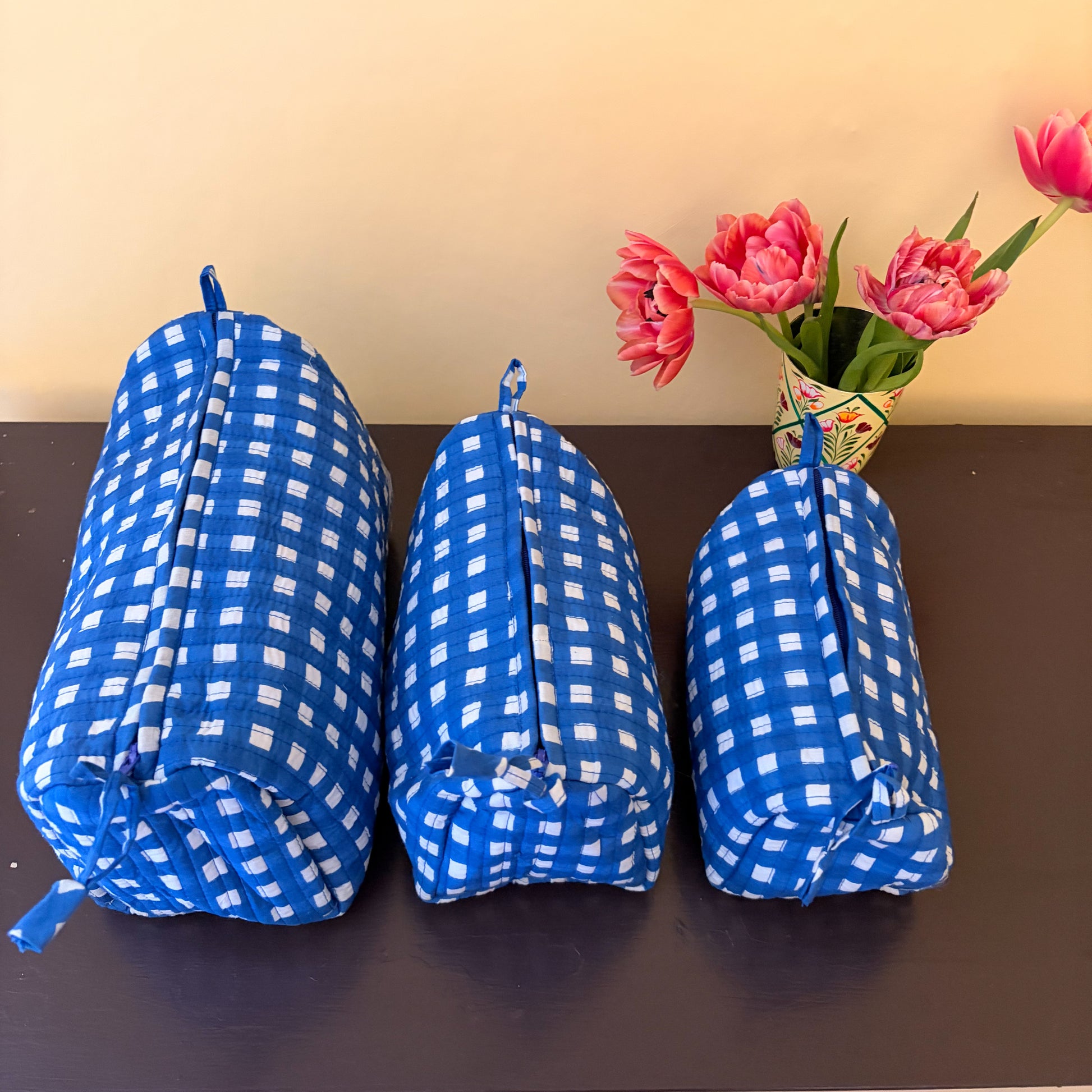Three blue and white checkered cosmetic bags on a dark surface with pink flowers in the background.