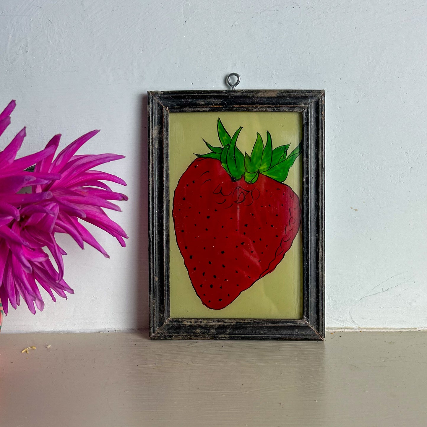 Framed artwork of a red strawberry on a light surface with a pink flower in the background.