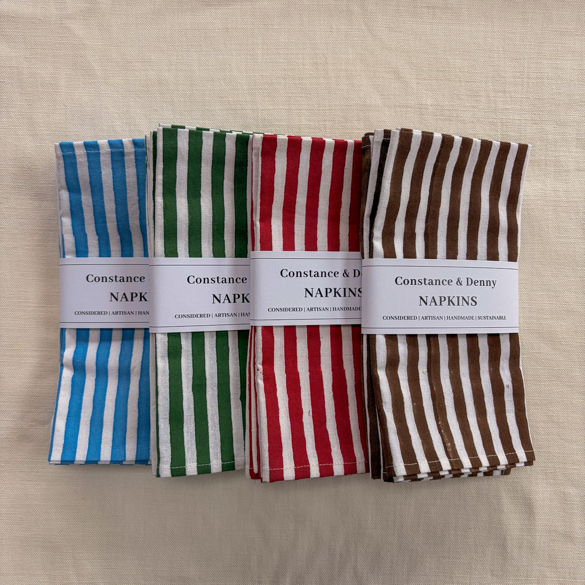 Four folded striped napkins in blue, green, red, and brown-and-white patterns, each wrapped with a white paper band labeled Constance Napkins, arranged on a beige fabric background.