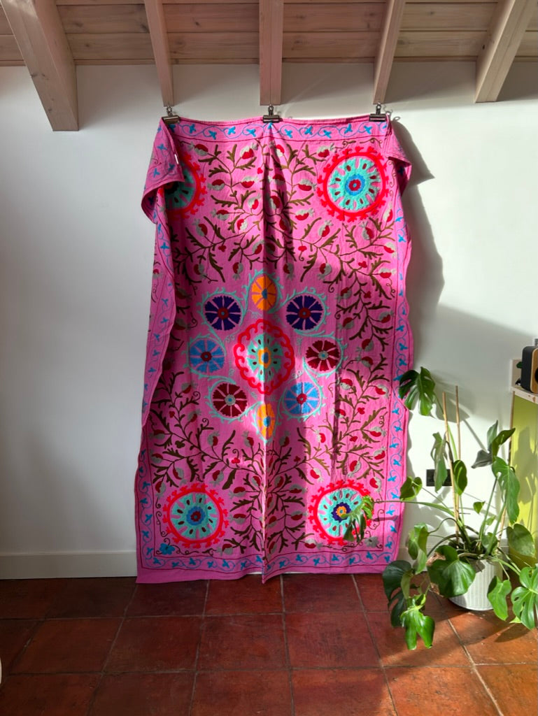 Pink suzani with colorful patterns hanging on a rack against a white wall.