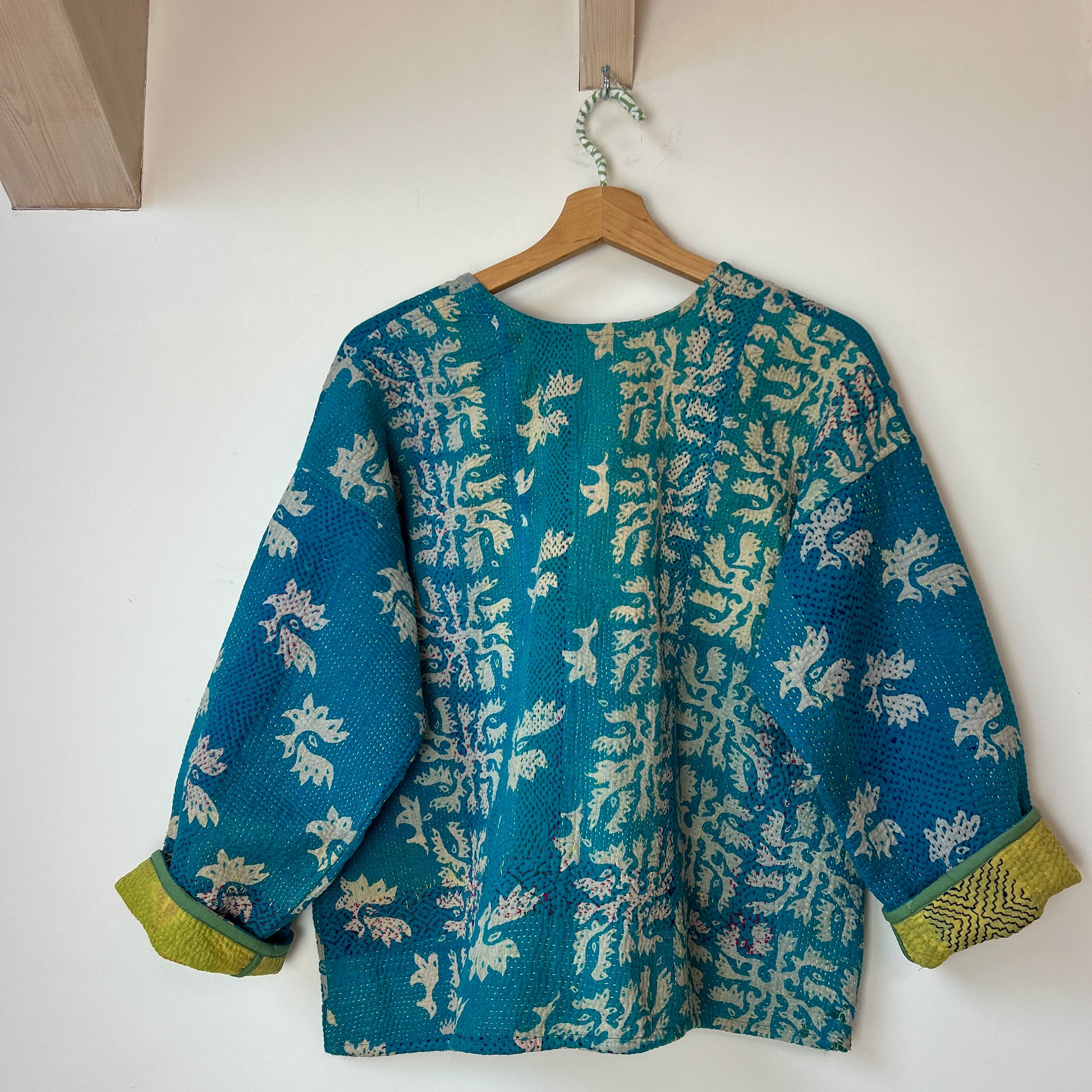 turquoise kantha with white floral pattern on a wooden hanger against a white wall. te floral patterns on a white background. Vintage-kantha-jacket-handmade-unique-38-turquoise-pattern-stitching-reverse