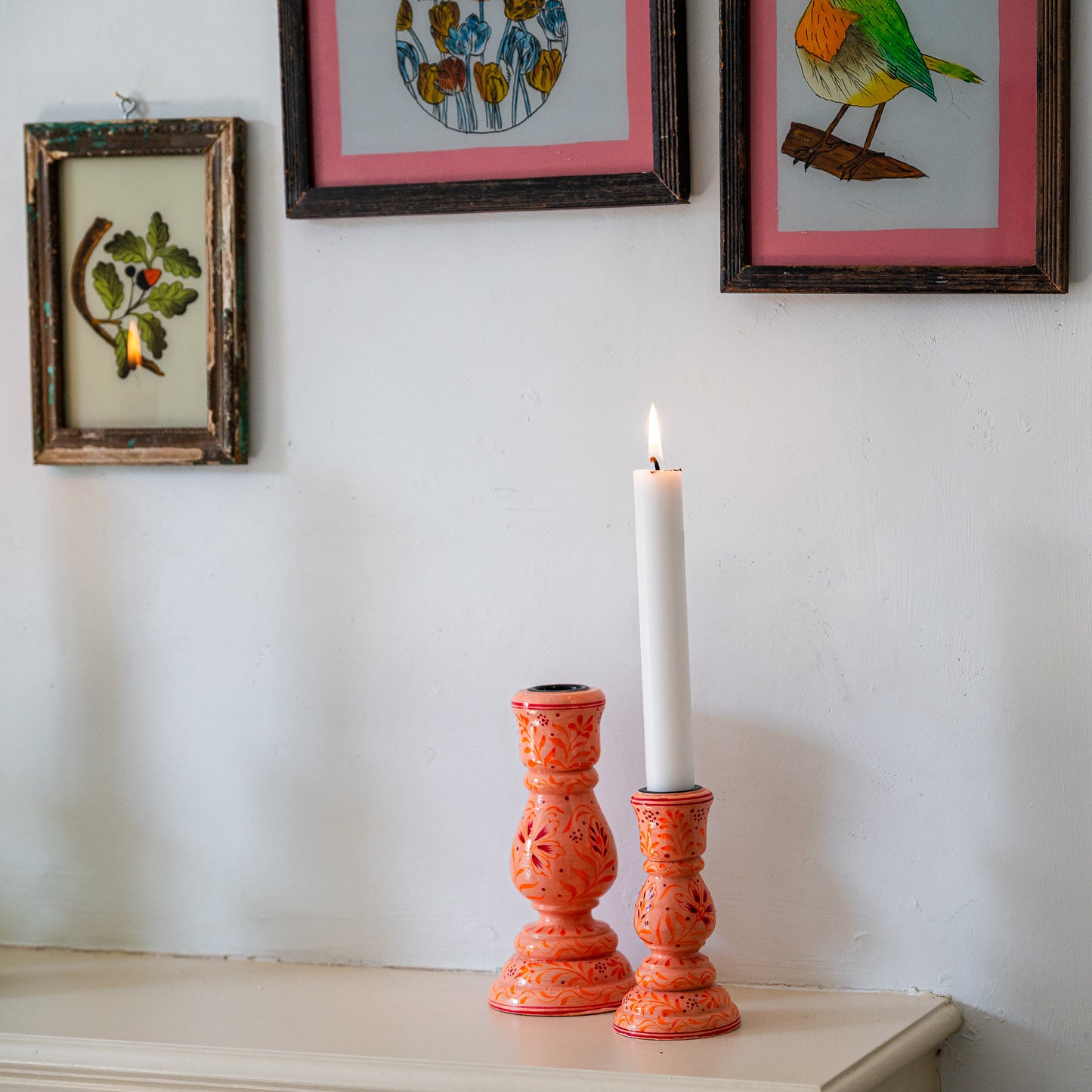 Two orange candle holders with a white candle on a white surface, framed artwork in the background.