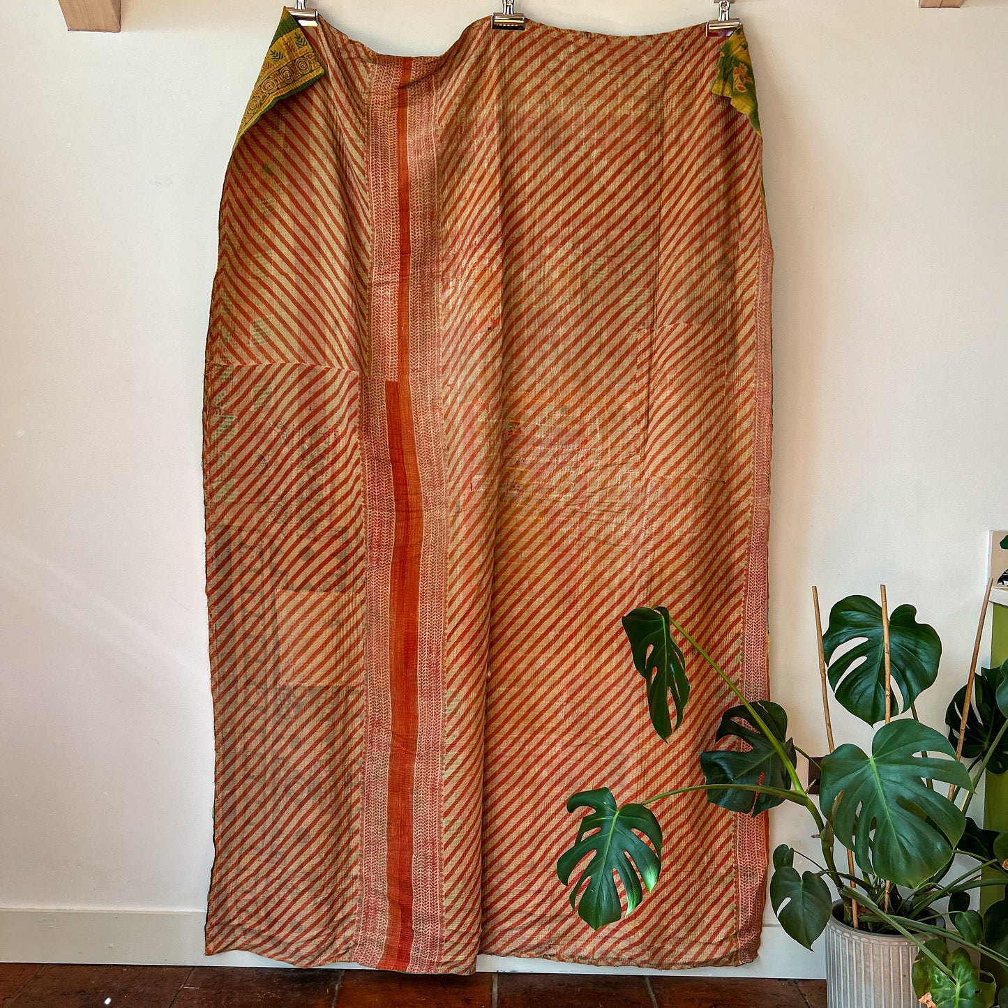 Long, patterned fabric hanging on a rod with a plant in the background. vintage-kantha-2015-textiles-blanket-rust-red