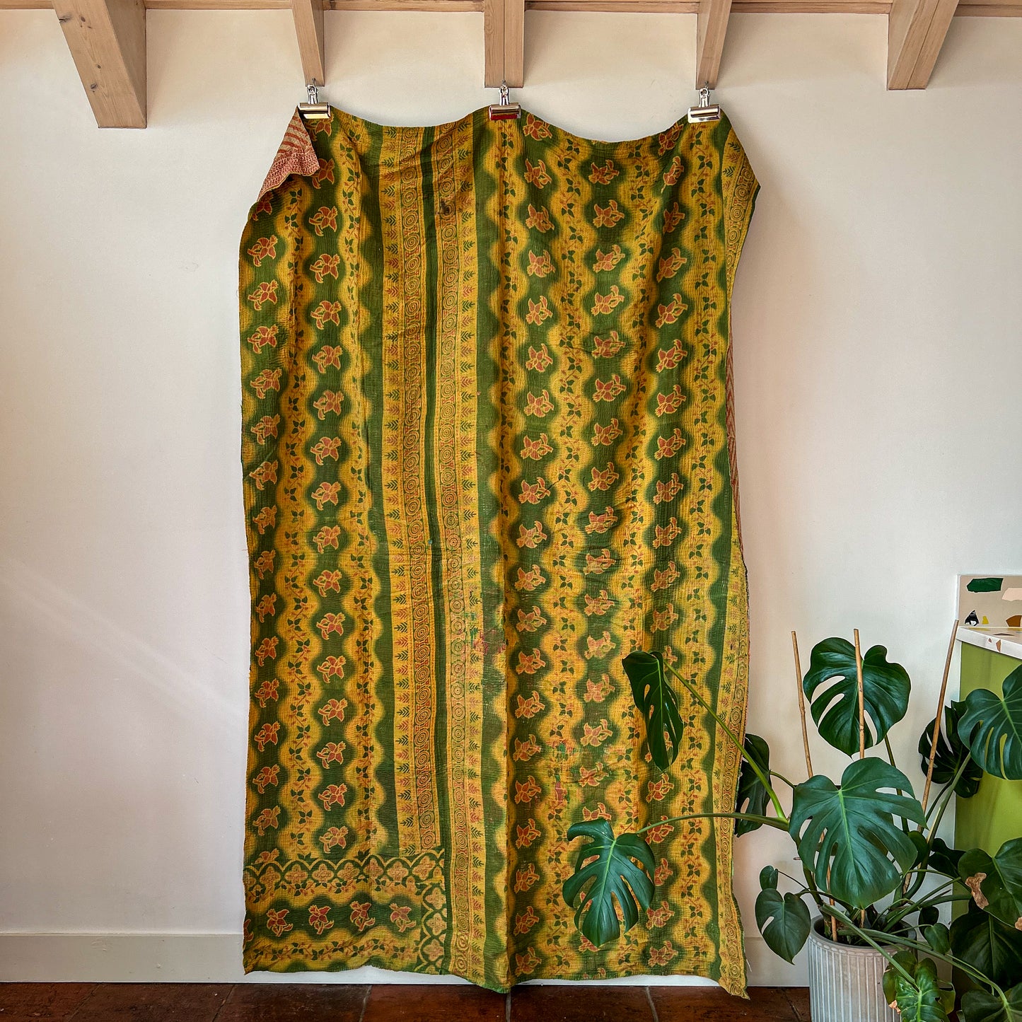 Green and yellow patterned dress hanging on a rack against a white wall with a plant in the corner.vintage-kantha-2015-textiles-blanket-green