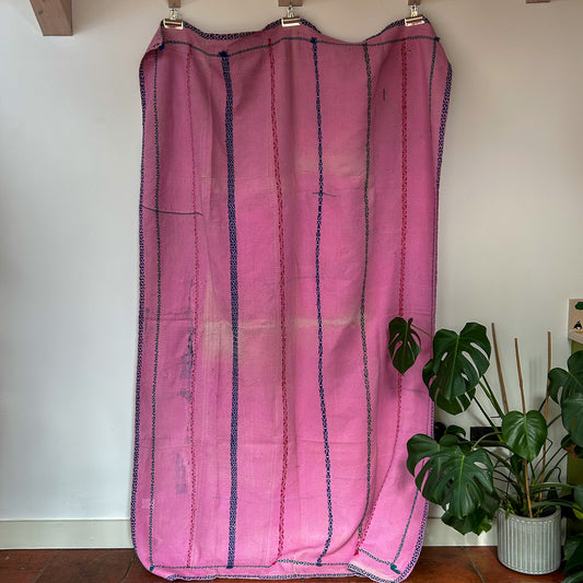 Pink fabric with blue stripes hanging on a rack against a white wall with plants in the background.