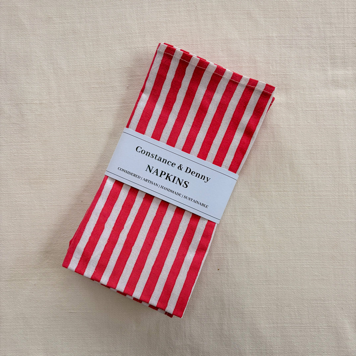 A folded stack of red and white striped paper napkins with a white paper label band reading 'Constance & Denny NAPKINS' on a beige linen background.