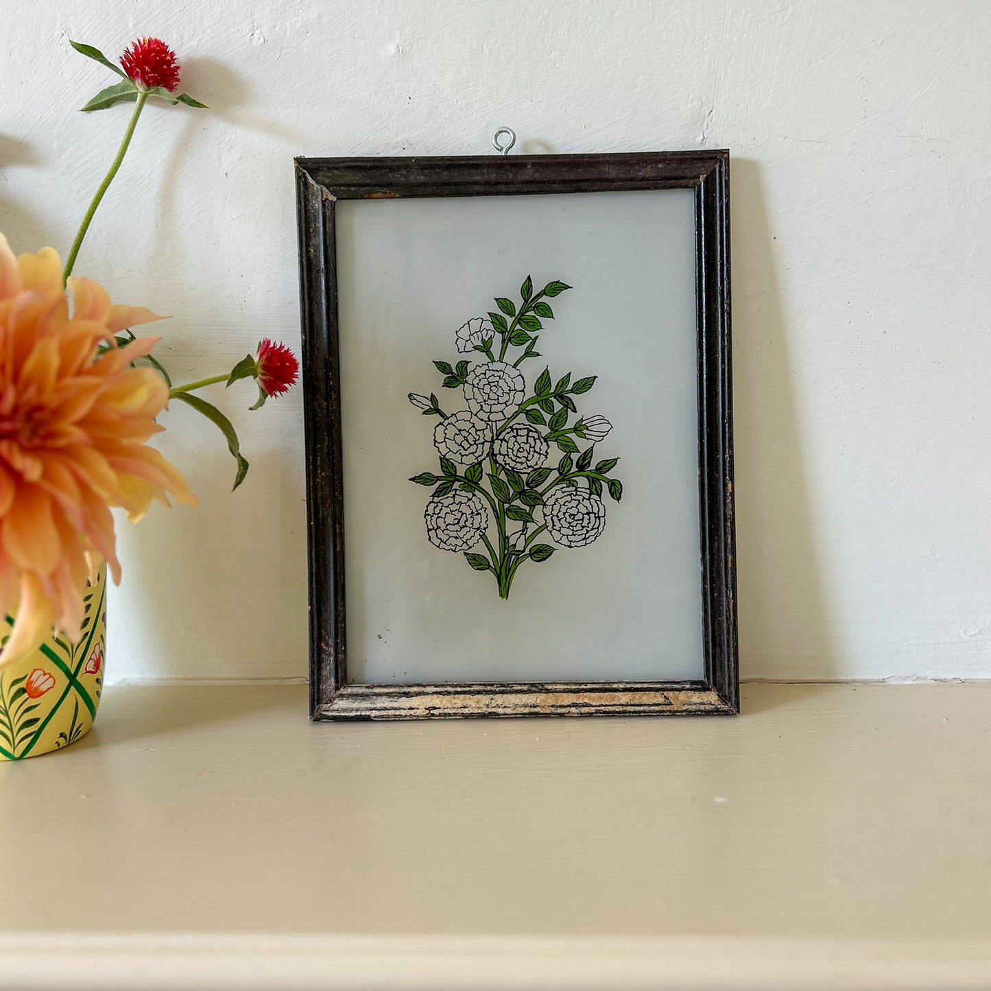 Framed floral artwork on a shelf with a vase of flowers. constance-and-denny-indian-reverse-glass-painting-boxed-packaging-hand-painted-artisan-art-white -carnations